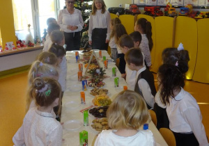 Pani dyrektor Maria Królikowska, pani Ewa Janicka oraz dzieci stoją wokół nakrytego białym obrusem i poczęstunkiem stołu.